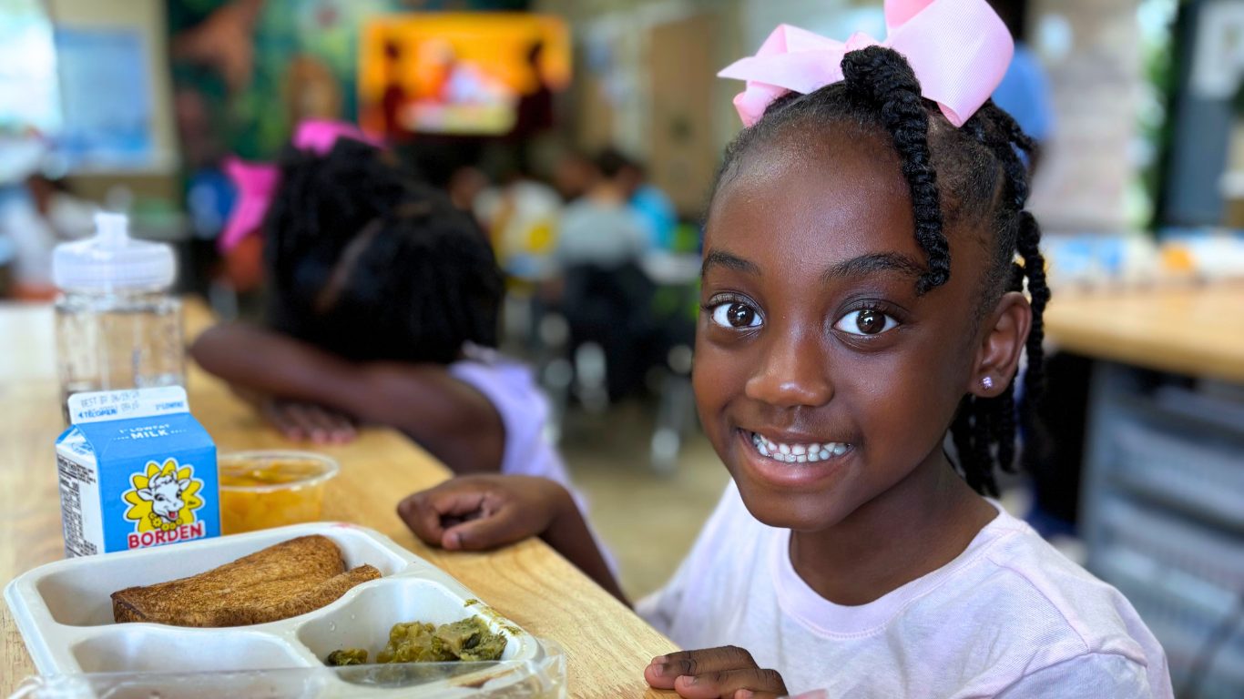 A smiling young girl with a pink bow sits at a school cafeteria table with a tray of toast, greens, and a carton of milk.